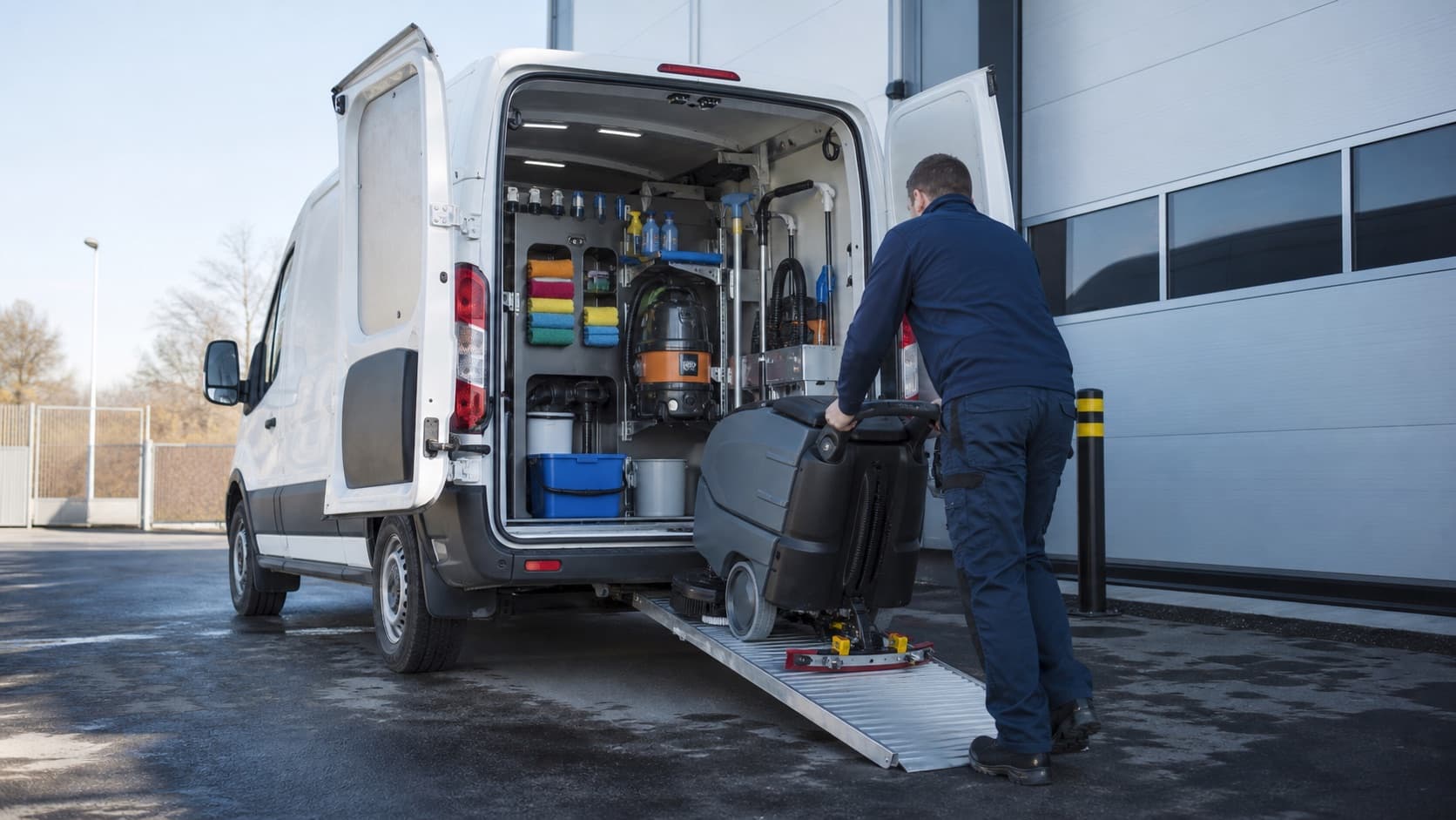 Medewerker rijdt een schrobzuigmachine via een oprijplaat uit een bestelwagen met geordend schoonmaakmaterieel.