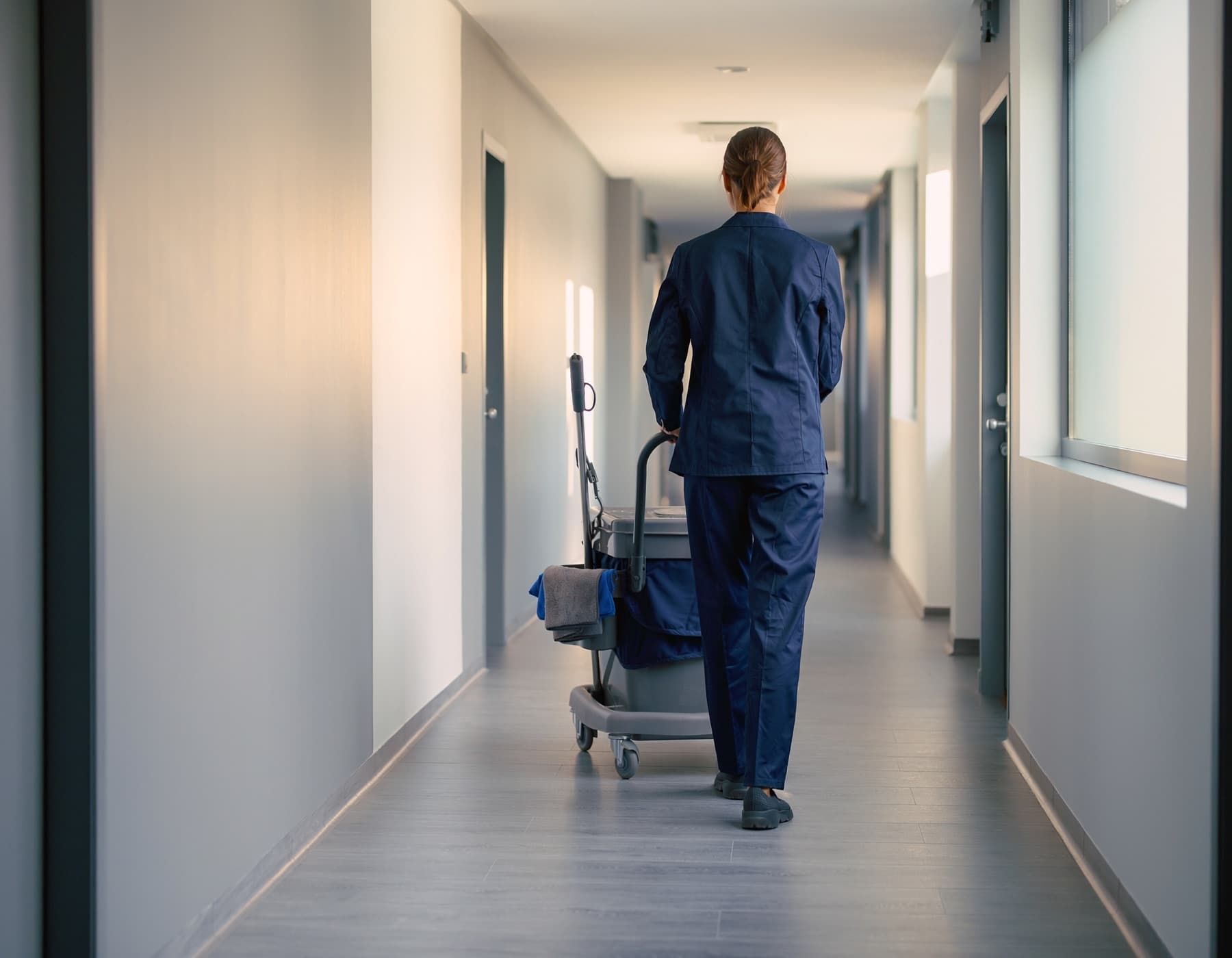 Medewerker in bedrijfskledij loopt met een schoonmaakkar door een rustige gang in een zorgomgeving.